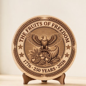 May include: A round wooden plaque with the text "THE FRUITS OF FREEDOM" and "1776 - 250 YEARS - 2026" encircling a central design. The design features an eagle, a cornucopia, and various fruits. The plaque is displayed on a small wooden stand.
