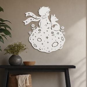 May include: A white metal wall art piece depicting the Little Prince and a fox sitting on a moon-shaped planet with stars and flowers. The artwork is mounted on a neutral-colored wall, with a black bench and decorative baskets in the foreground.