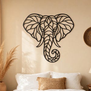 May include: A black, geometric elephant head wall art decor. The elephant's head is made of interconnected lines, creating a detailed, artistic design. The artwork is mounted on a beige wall above a bed with white linens and a brown throw.