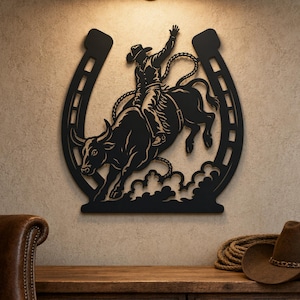 May include: A black metal wall art piece shaped like a horseshoe, featuring a silhouette of a cowboy riding a bucking bull. The design includes a rope trailing behind the rider and stylized clouds at the base, all rendered in a solid black finish.