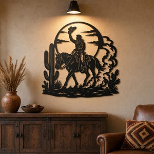 May include: Black metal wall art depicting a cowboy on horseback, raising his hat. The silhouette is set against a circular backdrop with clouds and a cactus, evoking a Western theme. The artwork is mounted on a neutral-toned wall.
