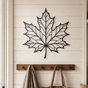 May include: A black metal maple leaf wall art decor. The leaf is mounted on a white wall with horizontal wood paneling. Below the leaf is a wooden coat rack with hooks, a beige scarf, and a woven tote bag.