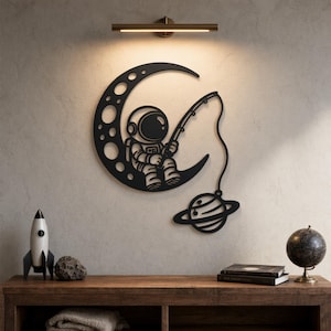 May include: A black metal wall art piece featuring an astronaut sitting on a crescent moon with craters, holding a fishing rod that extends downward to catch a small planet with rings. The design is flat, silhouette-style, and mounted on a wall.