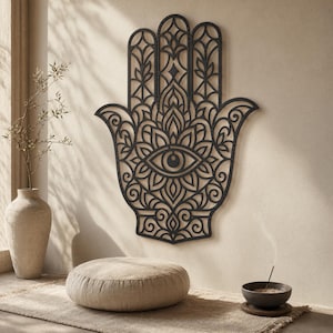 Peut inclure: Une décoration murale en bois noir en forme de main de Hamsa, finement sculptée. Le motif comprend un œil au centre et des motifs détaillés. L'objet est accroché sur un mur de couleur neutre, idéal pour la décoration intérieure.