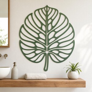 May include: A large, green, leaf-shaped wall decoration with intricate cut-out details. The leaf is mounted on a white wall above a wooden shelf. A small potted plant, a rolled towel, and a soap dispenser are on the shelf.