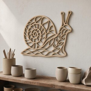 May include: A wooden snail wall art piece with a detailed spiral shell and leaf accents. The light brown art is mounted on a neutral wall. Below, a wooden shelf displays several beige ceramic cups and a cylindrical container with tools.