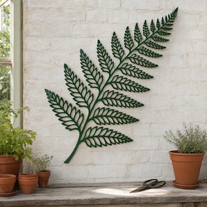 Può includere: Una grande decorazione da parete a forma di foglia di felce in metallo verde scuro, montata su un muro di mattoni bianchi. La felce presenta intricati motivi ritagliati. Lo sfondo include piante in vaso e una superficie in legno.