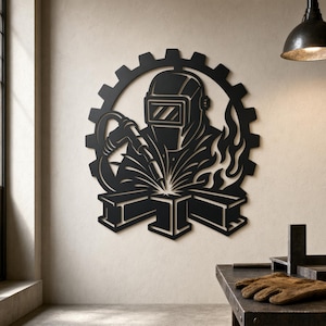 May include: A black metal wall art piece featuring a welder wearing a protective helmet and holding a welding torch. The design includes a circular gear frame, stylized flames, and structural steel beams at the base, creating a detailed silhouette.