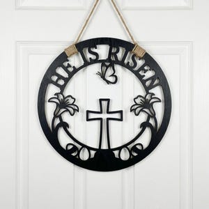 Könnte beinhalten: Schwarzes hölzernes Ostertürschild mit den Worten "HE IS RISEN" in einem Kreis ausgeschnitten. Das Schild zeigt ein Kreuz, einen Schmetterling und florale Akzente. Es hängt an einem Naturseil.