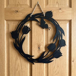 Puede incluir: Una corona de madera negra con diseño de tulipanes y mariposa. La corona está suspendida por un cordón de hilo natural. La corona está montada en una puerta de madera.