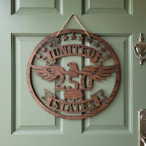 May include: A round wooden sign with the words "UNITED 250 STATES" cut out. The sign features an eagle with outstretched wings and star cutouts. It hangs from a natural twine string.