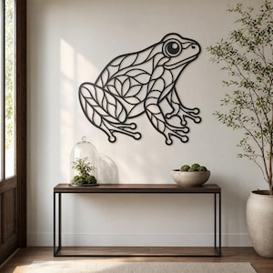 Puede incluir: Una obra de arte mural de metal negro con forma de rana con intrincados patrones en forma de hojas. La rana está montada en una pared blanca sobre una consola de madera oscura. La mesa tiene un marco de metal negro y una campana de cristal.