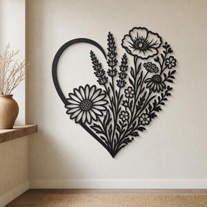 May include: Black metal wall art in the shape of a heart, filled with detailed floral designs. The artwork features a large daisy, poppy, and other flowers, creating a nature-inspired decorative piece. The heart shape is open, with a curved top.