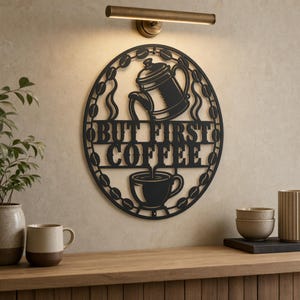 Op de afbeelding: Een rond zwart metalen wandbord met een uitgesneden ontwerp van een cafetière die in een kopje schenkt, omringd door een rand van koffiebonen. De tekst "BUT FIRST COFFEE" staat in het midden in een vetgedrukt schreeflettertype.