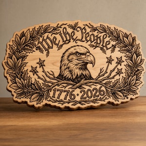 May include: Wooden plaque with an engraved design featuring an eagle's head, the words "We the People," and the dates "1776-2026." The design is surrounded by a wreath of leaves and stars. The plaque is made of light-colored wood.