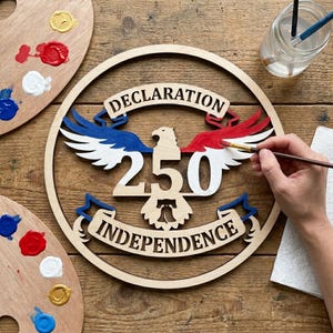 May include: A wooden circular sign with the text "DECLARATION INDEPENDENCE" and "250" in the center. The design includes an eagle and wings painted in red, white, and blue. Paint palettes and a paintbrush are also visible.