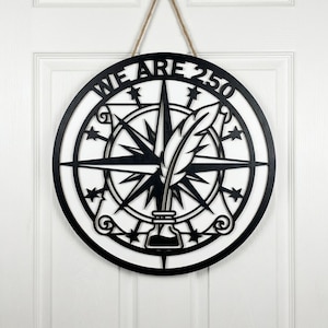 May include: A round, black and white wooden sign with the text "WE ARE 250". The sign features a compass rose design with a quill and inkwell in the center. It hangs from a natural twine cord.