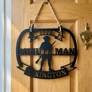 May include: Black metal sign with the words "1775", "Minute Man", and "Lexington" cut out. The sign features a silhouette of a colonial soldier holding a rifle. The sign is suspended from a wooden door with a rope.