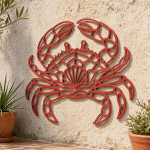 May include: A red crab wall art piece with a detailed, openwork design. The crab has large claws, multiple legs, and a shell-like pattern on its body. The art is mounted on a textured, light-colored wall, with potted plants in the foreground.