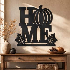 May include: Black metal wall art spelling "HOME" with a pumpkin replacing the "O". Two leaves are at the bottom. The art is mounted on a beige wall above a wooden table with autumn decor.