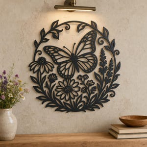 May include: A circular black metal wall art piece featuring a detailed butterfly at the center, surrounded by a wreath of various flowers, leaves, and stems. The design is intricate with cut-out patterns, creating a silhouette effect against the wall.