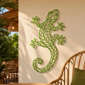 May include: A green metal lizard wall decoration. The lizard features a leaf-like pattern and is mounted on a light-coloured wall. A wicker chair and greenery are visible in the background, creating a tropical feel.