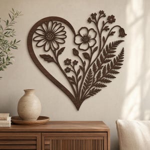 Può includere: Un'opera d'arte da parete in legno marrone scuro a forma di cuore, con disegni floreali. Il cuore è riempito con una margherita, un fiore e foglie simili a felci. L'opera è montata su una parete beige chiaro.