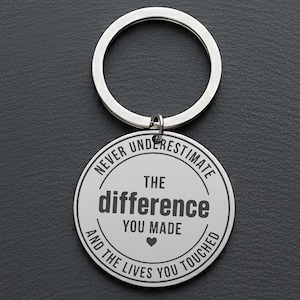 May include: Silver-colored keychain with a round pendant. The pendant has the text "NEVER UNDERESTIMATE THE difference YOU MADE and THE LIVES YOU TOUCHED" in black, with a heart symbol. The keychain ring is also silver.