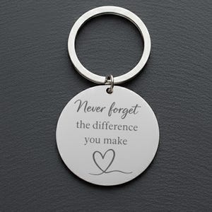 May include: A silver-colored keychain with a round pendant. The pendant has the engraved text "Never forget the difference you make" with a heart symbol below. The keychain ring is attached to the pendant with a small loop.