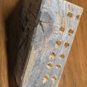 Puede incluir: Un bloque de madera rectangular y envejecido, de color gris-marrón natural. La veta de la madera es visible y el bloque tiene varios agujeros pequeños y espaciados uniformemente. Probablemente sea un objeto decorativo o funcional.