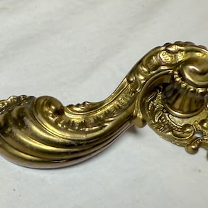 May include: Two antique brass door handles with ornate, swirling designs. The handles have a curved shape and are likely from a vintage or classic style door. The brass has a warm, aged patina, suggesting they are old.