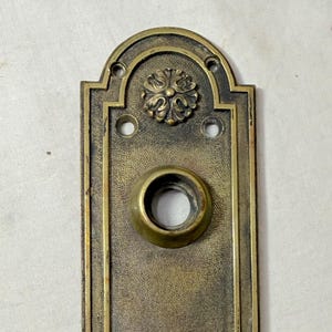 May include: Antique brass door plate with decorative floral accents at the top and bottom. The plate has a rectangular shape with a central hole for a doorknob and a smaller hole below.