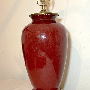 May include: A red ceramic lamp with a brass base and a gold-tone lampshade. The lamp has a classic design and would be a great addition to any home.