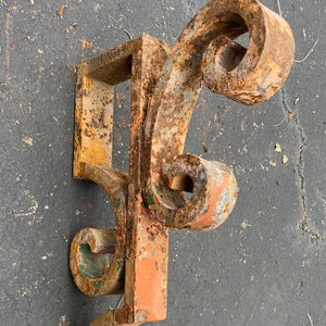 May include: An antique, ornate metal bracket with a weathered, rusty finish. The bracket features a rectangular base and decorative scrollwork with remnants of orange, green, and yellow paint. A vintage architectural element.