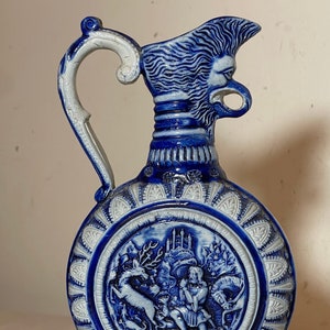May include: A blue and white ceramic pitcher with a detailed design. The pitcher features a round body with a raised, circular scene of a deer and a person in a forest. The pitcher has a handle shaped like a lion's head and a base with a blue and white design.