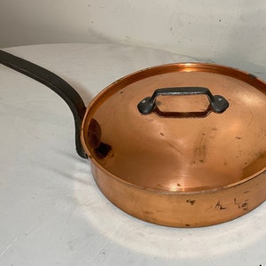 May include: A copper pot with a black iron handle and lid. The pot has a rounded bottom and a wide opening. The lid has a small handle on top.