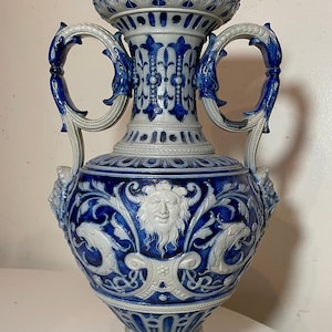 May include: A blue and white ceramic vase with two handles in the shape of dragons. The vase is decorated with a grotesque face and floral designs.