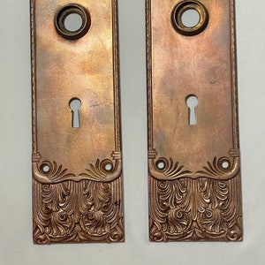 May include: A pair of antique brass door plates with ornate floral designs. The plates have a keyhole and a round hole for a doorknob.