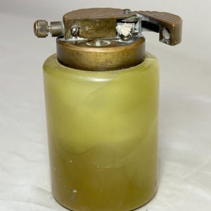 May include: Vintage lighter with a cylindrical, olive-green body and a brass top. The lighter features a hinged lid and a textured design on the top. The lighter is a classic collectible item.