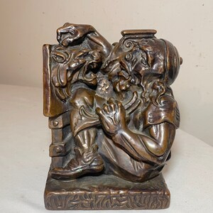 Puede incluir: Un sujetalibros de bronce que representa a un hombre luchando contra una bestia m&iacute;tica. El hombre lleva un casco y una t&uacute;nica, y la bestia tiene una cara aullante y garras. El sujetalibros es detallado y tiene una base texturizada.
