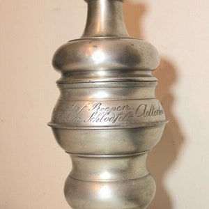 May include: A silver pewter container with a pointed top and a base. The container has an inscription that reads "Letlef. Breeper, Allerheiligen Schloofelt".