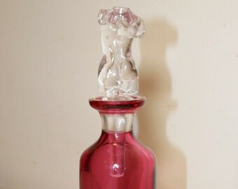 May include: A clear glass bottle stopper with a sculpted human torso design. The bottle is a deep burgundy color with a clear glass base.