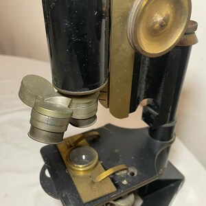May include: An antique microscope with a black body and brass components. The microscope features multiple lenses, adjustment knobs, and a stage for slides. The design suggests a vintage scientific instrument, likely used for biological or medical research.