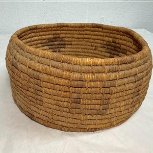 May include: A round, coiled basket constructed from natural brown fibers. The basket features a consistent, layered weave pattern with visible vertical stitching binding the horizontal coils together, creating a sturdy, open-top cylindrical container.