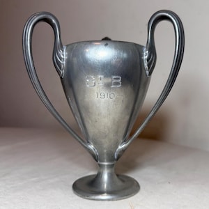 May include: A silver-toned trophy cup with two curved handles. The cup has a flared shape and a pedestal base. The inscription "S B" and the year "1910" are etched on the front. The cup is likely vintage.