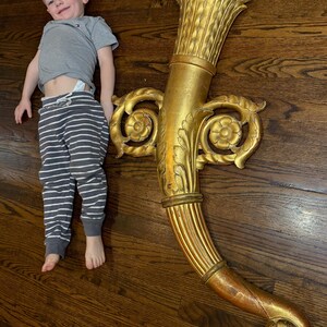 May include: A large, ornate, gold-colored decorative wall sconce with a curved design. The sconce features intricate floral and scrollwork details. The image also includes a child wearing a gray t-shirt and striped pants.
