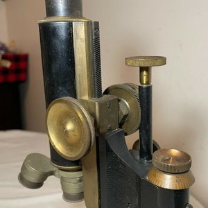 May include: An antique microscope with a black body and brass components. The microscope features a series of adjustment knobs and lenses, with a focus on the intricate details of its design. The base is black, and the overall aesthetic is vintage.