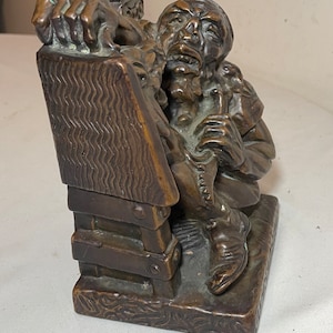 Puede incluir: Una escultura de bronce de un hombre sentado en una silla de madera, sosteniendo una pipa en su mano. La escultura es detallada y realista, con una superficie texturizada.