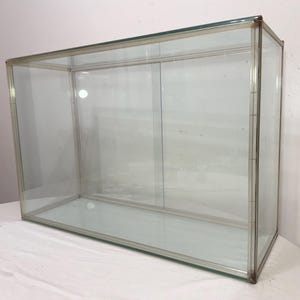 May include: A rectangular glass display case with a silver metal frame. The case is empty and has a sliding glass door. The glass is clear, allowing full visibility of the interior. Ideal for showcasing collectibles.
