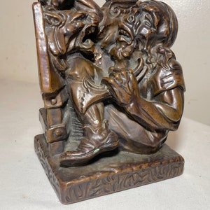 Puede incluir: Un sujetalibros de bronce que representa a un hombre siendo atacado por una g&aacute;rgola. El hombre lleva una t&uacute;nica y un turbante, y la g&aacute;rgola le est&aacute; ara&ntilde;ando la cara. El sujetalibros est&aacute; sobre una base cuadrada con un dise&ntilde;o decorativo.